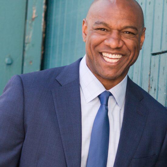 A bald Black man wearing a blue suit and tie smiles for a photograph
